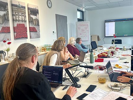 Mehrere Teilnehmende aus den Ortsvereinen sitzen an einer langen Tischtafel und schauen auf die Beamerleinwand, auf die der Kursleiter die Informationen projeziert