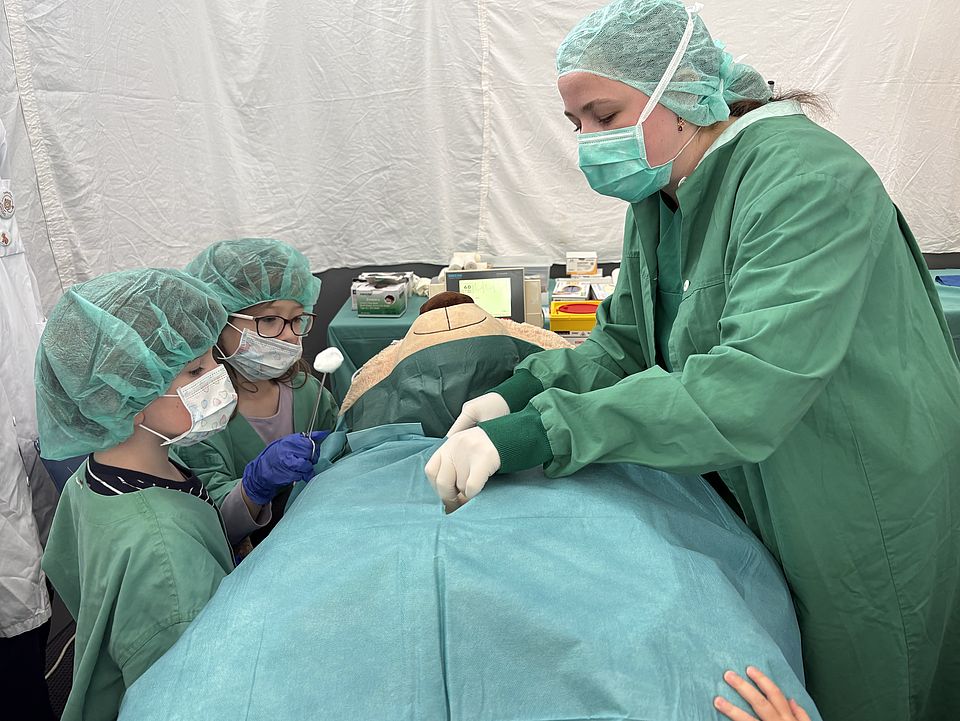2026_06_01TBK_Teddybärkrankenhaus_UMG__Kita_Bovenden_8_.jpg Mehrere Kinder tragen OP-Hauben und Schutzkleidung und stehen zusammen mit einer betreuenden Person an einem Tisch, auf dem ein Teddybär unter einem grünen Tuch liegt. Der Teddy wird spielerisch „operiert“.