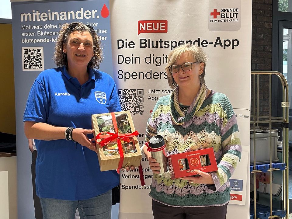 2026_09_25_Blutspende_Dassel__6_.jpeg Zwei Personen stehen vor einem Informationsstand des DRK-Blutspendedienstes. Die geehrte Blutspenderin erhält Präsente anlässlich ihres Blutspendejubiläums.
