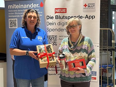 Zwei Personen stehen vor einem Informationsstand des DRK-Blutspendedienstes. Die geehrte Blutspenderin erhält Präsente anlässlich ihres Blutspendejubiläums.