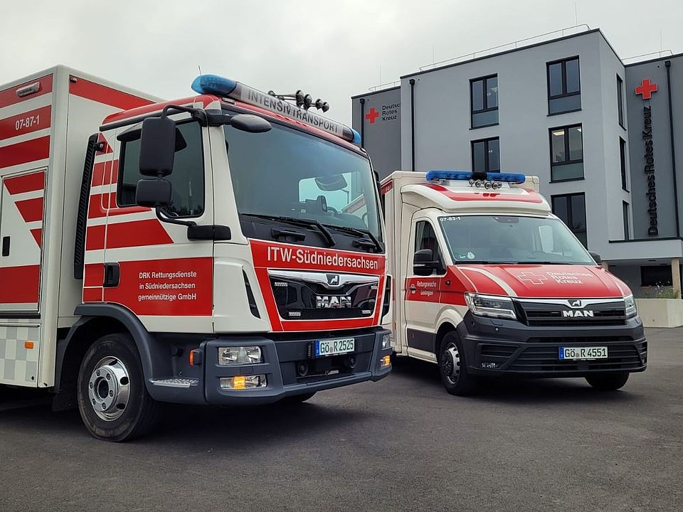 Vor dem modernen, mehrstöckigen Gebäude des Deutschen Roten Kreuzes in Göttingen stehen zwei Einsatzfahrzeuge. Links befindet sich ein großer Intensivtransportwagen (ITW) mit rot‑weißer Beschriftung „ITW Südniedersachsen“. Rechts daneben steht ein Rettungswagen des DRK in gleicher Farbgebung. Beide Fahrzeuge sind frontal sichtbar und stehen auf einem asphaltierten Vorplatz. Am Gebäude im Hintergrund ist das rote DRK‑Logo sowie die Beschriftung „Deutsches Rotes Kreuz“ erkennbar. Der Himmel ist bedeckt, und die Szene wirkt ruhig und geordnet.