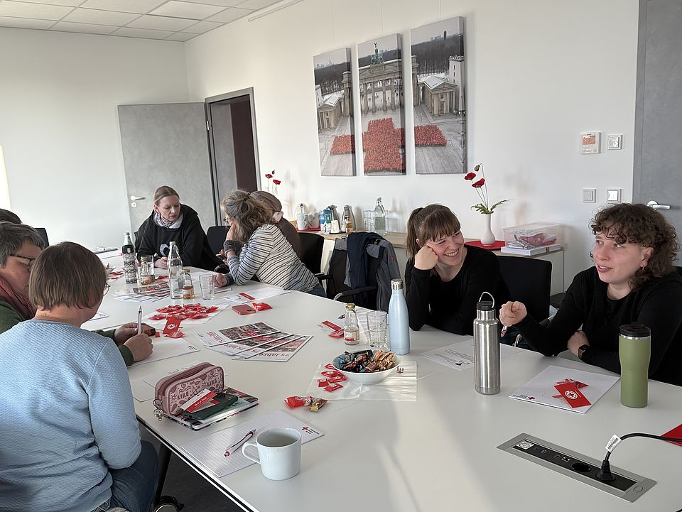 Ein heller Konferenzraum des Deutschen Roten Kreuzes in der Zimmermannstraße 4 in Göttingen. Mehrere Personen sitzen an einem großen weißen Konferenztisch und nehmen an der Einführungsveranstaltung für neue Schulbegleiterinnen des FUD im DRK‑Kreisverband Göttingen‑Northeim e. V. teil. Auf dem Tisch liegen Schulungsunterlagen, DRK‑Infomaterialien, kleine rote Give‑aways, Getränke und eine Schale mit Süßigkeiten. Im Hintergrund steht ein Sideboard mit weiteren Getränken und Materialien. An der Wand hängen drei große Bildtafeln, die ein Motiv des Brandenburger Tors mit einem roten Teppich zeigen. Die Teilnehmenden arbeiten konzentriert, schreiben Notizen oder sprechen miteinander. Die Schulung wird von Alexandra Jäger geleitet.