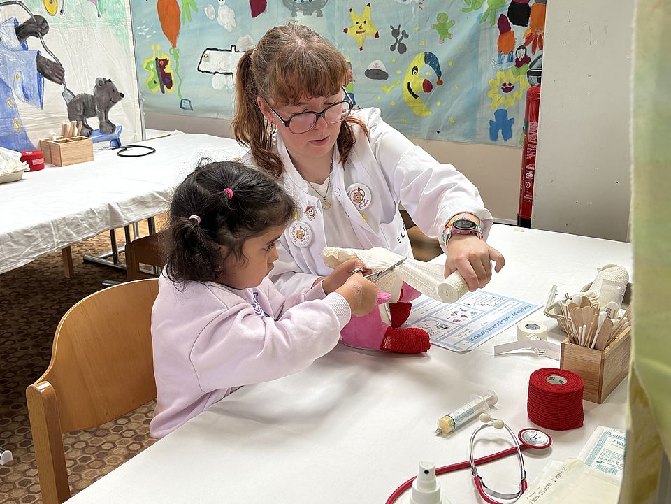 2026_06_01TBK_Teddybärkrankenhaus_UMG__Kita_Bovenden_10_.jpg An einem Tisch sitzt ein Kind und verbindet mit Anleitung einer betreuenden Person ein Kuscheltier. Medizinische Utensilien wie Verbände und ein Stethoskop liegen bereit, im Hintergrund hängen bunte Kinderzeichnungen.