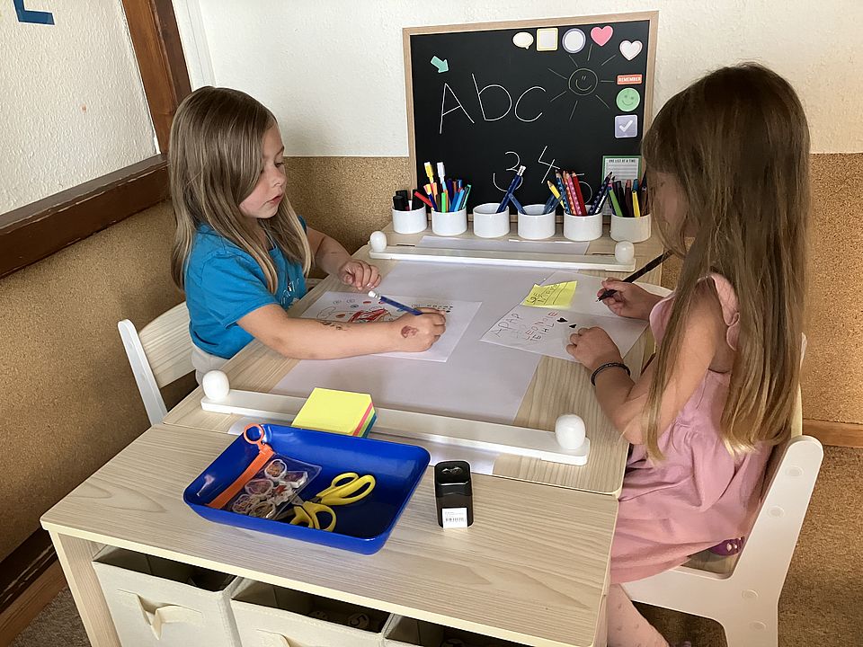 zwei Mädchen sitzen an einem Tisch und bemalen ein Blatt Papier. An der Seite lehnt eine Schultafel an der Wand, an der ABC steht und davor stehen Stifte in Bechern