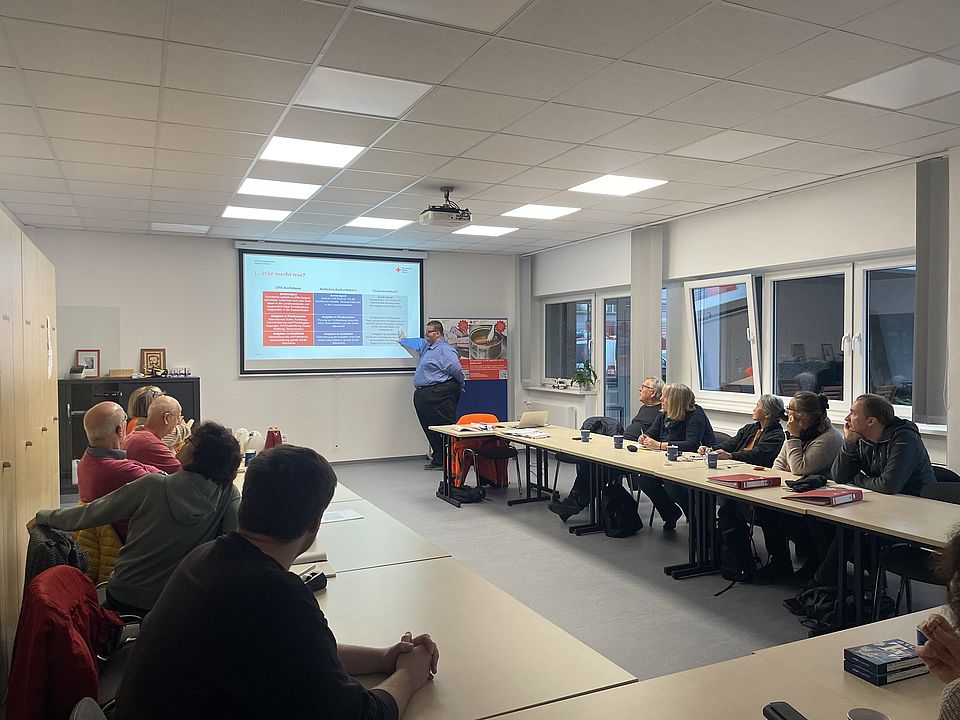 Teilnehmende der Schulung sitzen an Tischen und hören dem Vortragenden zu, der an einer Beamer Leinwand eine Tabelle erläutert