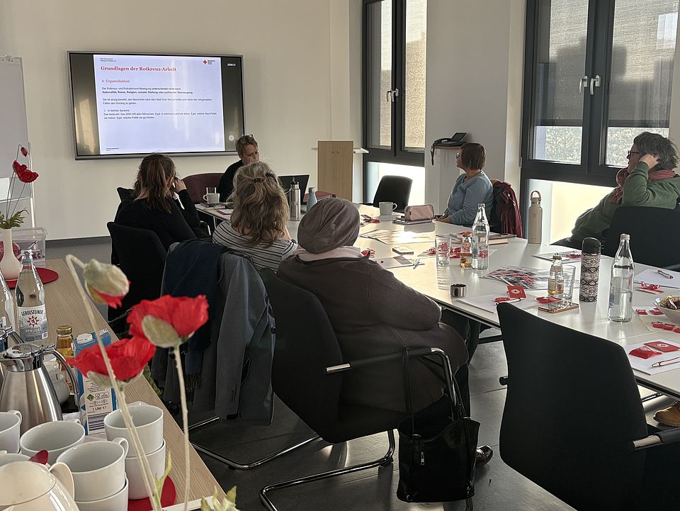 Ein heller Schulungsraum mit mehreren Personen, die an einem großen rechteckigen Tisch sitzen und an einer Einführungsveranstaltung für neue Schulbegleiterinnen des FUD im DRK‑Kreisverband Göttingen‑Northeim e. V. teilnehmen. Auf einem großen Bildschirm an der Wand ist eine Präsentationsfolie zu sehen, auf die die Gruppe blickt. Auf dem Tisch liegen Unterlagen, Schreibmaterialien und Informationsflyer des DRK; außerdem stehen dort Wasserflaschen, Kaffeekannen, Tassen sowie einige dekorative rote Papierblumen. Die Schulung wird von Alexandra Jäger geleitet. Durch mehrere große Fenster fällt Tageslicht in den Raum in der Zimmermannstraße 4 in Göttingen