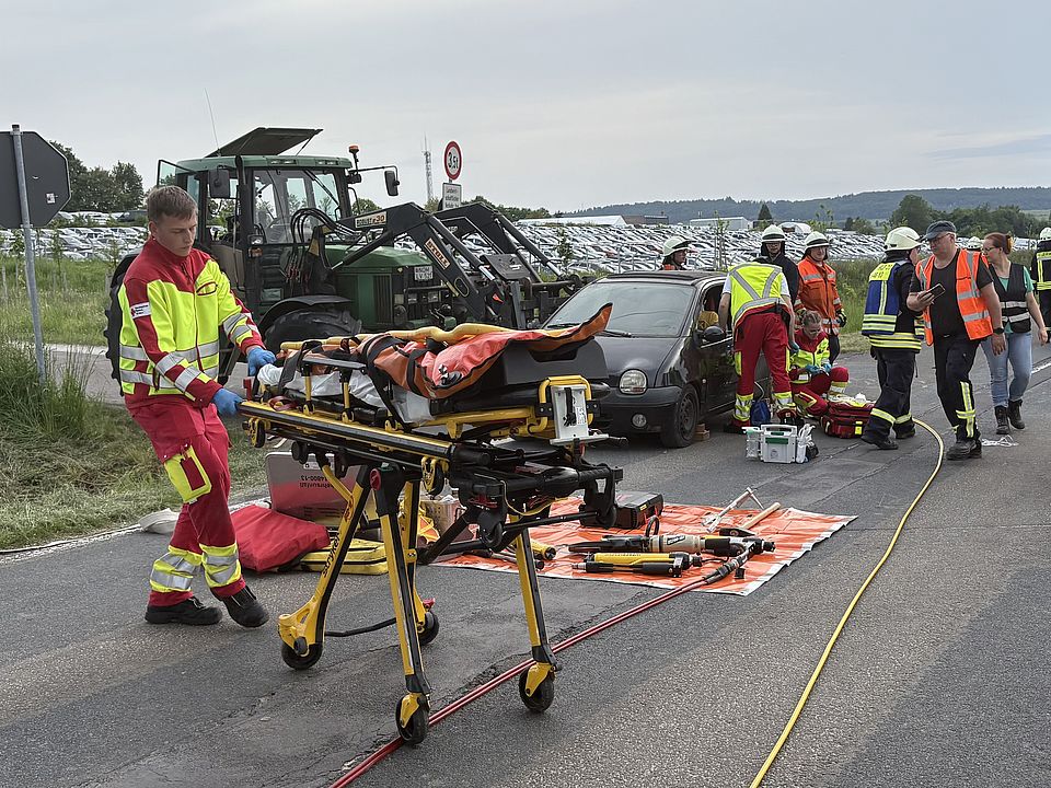 Rettungsdienstübung Uslar: Auszubildender des DRK-Rettungsdienstes in rot-gelber Uniform mit Patientenliege, im Hintergrund der Unfall mit Feuerwehrmitarbeitenden und Rettungsdienst