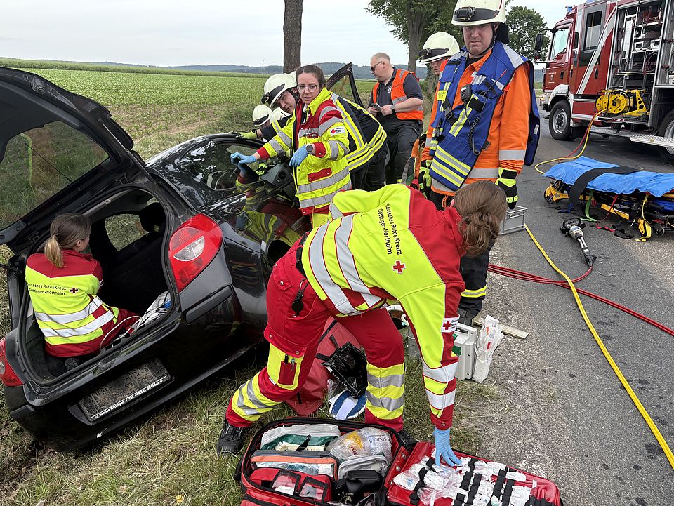 Rettungsdienst-Übung in Uslar: Drei Auszubildende des DRK-Rettungsdienstes vor einem Auto im Graben, eine Person ist im offenen Kofferraum des Autos, eine holt etwas aus einer Medizintasche, herum stehen Einsatzkräfte der Feuerwehr.