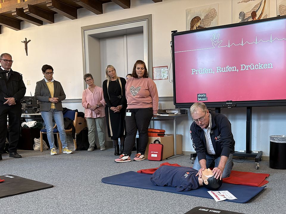 2026_09_21_Markus_Schiffer_demonstriert_wichtige_Handgriffe.jpg Im Rahmen der Veranstaltung erklärt der Fachbereichsleiter Erste Hilfe die Wiederbelebung anhand eines Reanimationsphantoms. Schüler*innen und Gäste verfolgen die Demonstration. Auf dem Bildschirm im Hintergrund sind die Kernschritte „Prüfen, Rufen, Drücken“ zu sehen, die die wichtigsten Maßnahmen im Notfall veranschaulichen