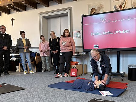 Im Rahmen der Veranstaltung erklärt der Fachbereichsleiter Erste Hilfe die Wiederbelebung anhand eines Reanimationsphantoms. Schüler*innen und Gäste verfolgen die Demonstration. Auf dem Bildschirm im Hintergrund sind die Kernschritte „Prüfen, Rufen, Drücken“ zu sehen, die die wichtigsten Maßnahmen im Notfall veranschaulichen