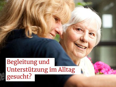 Ein Werbemotiv des Deutschen Roten Kreuzes (DRK) zeigt zwei Personen in einer Alltagssituation. Links im Bild sitzt eine Person, die anhand der Kleidung als Mitarbeitende des Familienunterstützenden Dienstes (FUD) des DRK erkennbar ist. Rechts daneben befindet sich eine weitere Person, die als Seniorin und Kundin des Dienstes dargestellt wird. Beide sitzen nah beieinander, und im Vordergrund sind farbige Blumen sichtbar. Im oberen Bereich des Bildes befindet sich das DRK‑Logo mit dem Schriftzug „Deutsches Rotes Kreuz“ und „DRK‑Kreisverband Göttingen‑Northeim e. V.“. Unterhalb des Motivs steht der Text: „Begleitung und Unterstützung im Alltag gesucht?“. Am unteren Rand befindet sich ein roter Balken mit der weißen Aufschrift: „Freie Kapazitäten beim Familienunterstützenden Dienst“.