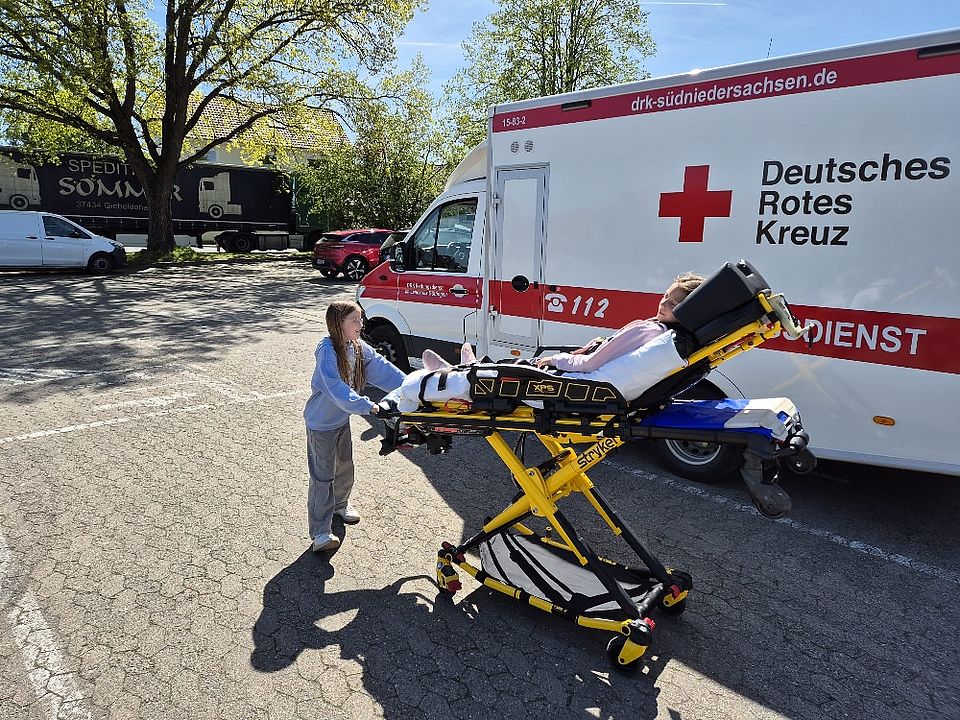 Ein mädchen schiebt eine Schülerin auf einer Trage liegend in Richtung eines Krankenwagens