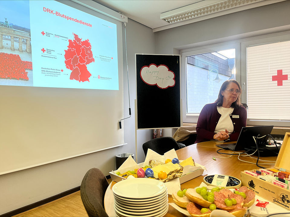 2026_03_12_Blutspendeworkshop__5_.jpeg An einem Tisch sitzt eine ehrenamtliche Mitarbeiterin des DRK-Kreisverbandes Göttingen Northeim an einem Laptop und hält einen Vortrag zur Blutspende, anlässlich eines Austauschworkshops zur Blutspende. Auf dem Tisch stehen Getränke und Arbeitsmaterialien und Essen und im Hintergrund ist ein rotes Kreuz am Fenster