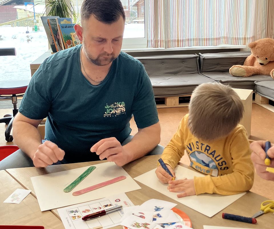 In einem Gruppenraum der DRK‑Kita Scheden sitzen eine erwachsene Begleitperson und ein Kind gemeinsam an einem Tisch. Vor ihnen liegen Bastelmaterialien, Holzstäbchen, Stifte und Papier mit Paw‑Patrol‑Motiven. Die erwachsene Person arbeitet an einem Projekt aus farbigen Holzstäbchen, während das Kind konzentriert auf ein eigenes Blatt zeichnet. Neben den Materialien liegen Scheren, Kleber und ein kleiner Teller mit weiteren Bastelteilen. Durch die großen Fenster im Hintergrund ist eine verschneite Außenlandschaft zu sehen. Die Szene zeigt eine der Olympiaden‑Stationen, bei der Kinder zusammen mit ihren Vätern, Großvätern oder Onkeln Aufgaben gestalten – passend zu einer Figur der Serie „Paw Patrol“.