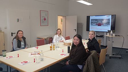Zukünftige Schulbegleiter*innen sitzen an einem Tisch im Konferenzraum des DRK in Hann. Münden, im Hintergrund ein Bildschirm mit DRK-Motiv und auf dem Tisch Getränke