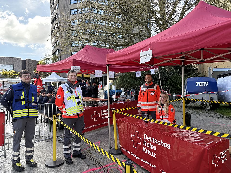 Helfer*innen und Mitglieder der DRK-Bereitschaft bei der Tour d'Energie 2026 in Göttingen beim Verpflegungsdienst vor einem Pavillion mit der Aufschrift 'Deutsches Rotes Kreuz' am Hiroshimaplatz