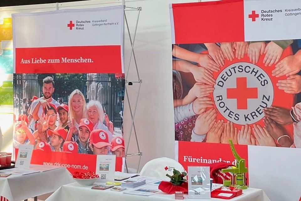 Ein Messestand des Deutschen Roten Kreuzes im Kreisverband Göttingen-Northeim e. V. auf der GöBit‑Ausbildungsmesse in Göttingen. Mehrere Infowände und Roll‑ups zeigen Fotos von DRK‑Einsatzkräften, das Rotkreuz‑Logo sowie Informationen zu verschiedenen Tätigkeitsbereichen. Auf Tischen liegen Flyer, Broschüren, Give‑aways und Informationsmaterial bereit. Links steht ein Glücksrad in Rot‑Weiß, das Besucherinnen und Besucher nutzen können. Rechts am Stand stehen mehrere DRK‑Mitarbeitende in Einsatzkleidung des Rettungsdiestes und sprechen mit interessierten Personen an einem Stehtisch. Der Stand ist hell beleuchtet und bietet vielfältige Informationen rund um Ausbildung, Ehrenamt und Aufgaben des DRK.