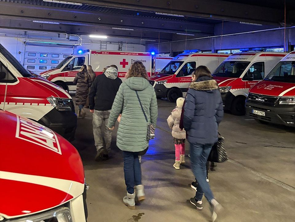 2026_01_22_in der Fahrzeughalle Man sieht eine Fahrzeughalle mit Rettungswagen und Fahrzeugen des DRK und eine Familie mit Erwachsenen und Kindern, die von hinten zu sehen sind und von der Kamera weglaufen, durch die Halle