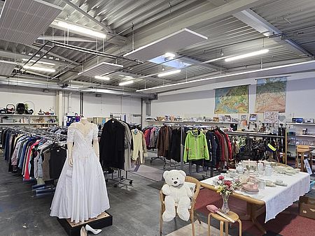 Man sieht das Göttinger Spendenzentrum des DRK von innen, mit vielen Ständern mit Kleidung und Tischen mit Gegenständen. Im Vordergrund sitzt ein weißer Teddybär und man sieht ein Hochzeitskleid an einem Mannequin