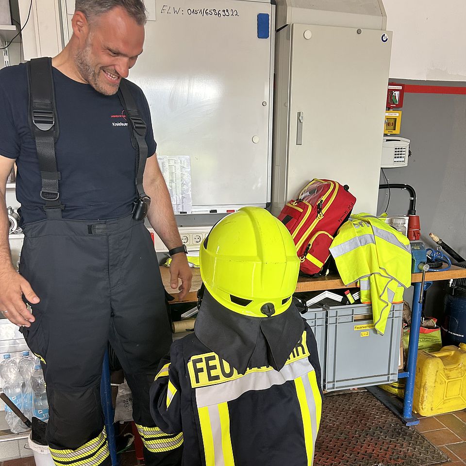 Kind probiert eine Feuerwehrjacke und Helm an, während eine Einsatzkraft der Feuerwehr Gimte daneben steht – Besuch der DRK-Kita Volkmarshausen.