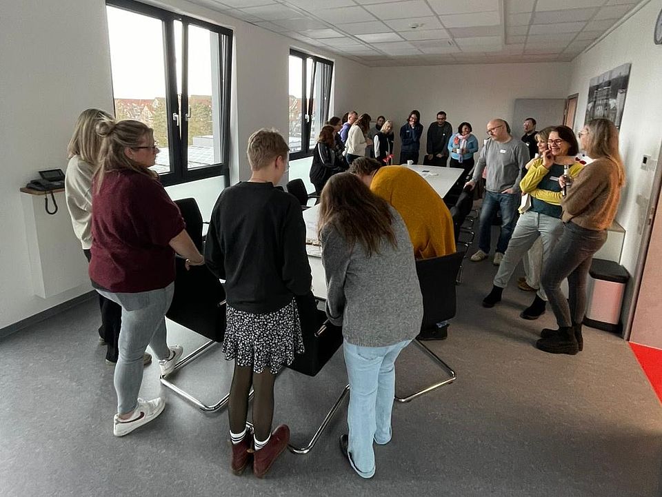 In einem Konferenzraum des DRK‑Kreisverbandes Göttingen‑Northeim e. V. steht eine große Gruppe von Teilnehmenden in einem Kreis um einen langen Konferenztisch. Auf dem Tisch liegen Arbeitskarten und Moderationsmaterialien. Der Raum ist hell, mit breiten Fenstern und einer Decke aus Akustikplatten. Die Teilnehmenden nehmen am Fachtag „Inklusiver Kinderschutz“ teil und bearbeiten im World‑Café‑Format Fragestellungen zur pädagogischen Haltung, zu Schutzkonzepten und zu Beschwerdemöglichkeiten für Kinder. Im Hintergrund stehen weitere Stühle und Tische sowie eine Präsentationsfläche.