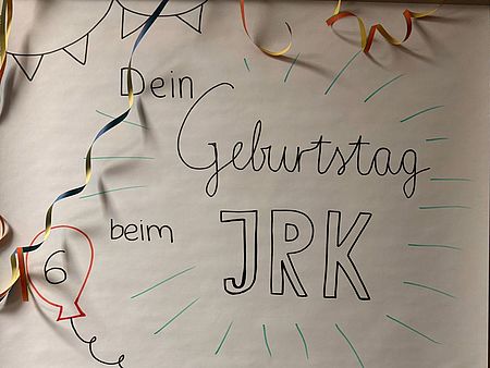 Man sieht ein Flipchart-Papier mit der Aufschrift "Dein Geburtstag beim JRK: Wunderversorgung, stabile Seitenlage, Blaulicht und...", sowie kleinen Ballons mit der Zahl 6 darauf und Luftschlangen darüberhängend