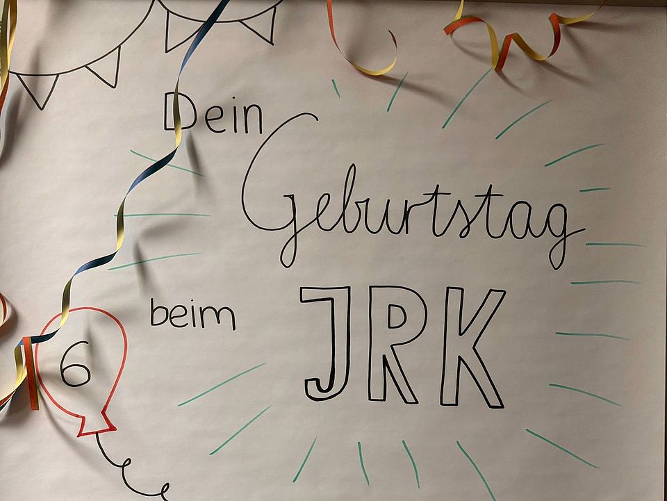 2026_01_22_Schild_Kindergeburtstag Man sieht ein Flipchart-Papier mit der Aufschrift "Dein Geburtstag beim JRK: Wunderversorgung, stabile Seitenlage, Blaulicht und...", sowie kleinen Ballons mit der Zahl 6 darauf und Luftschlangen darüberhängend
