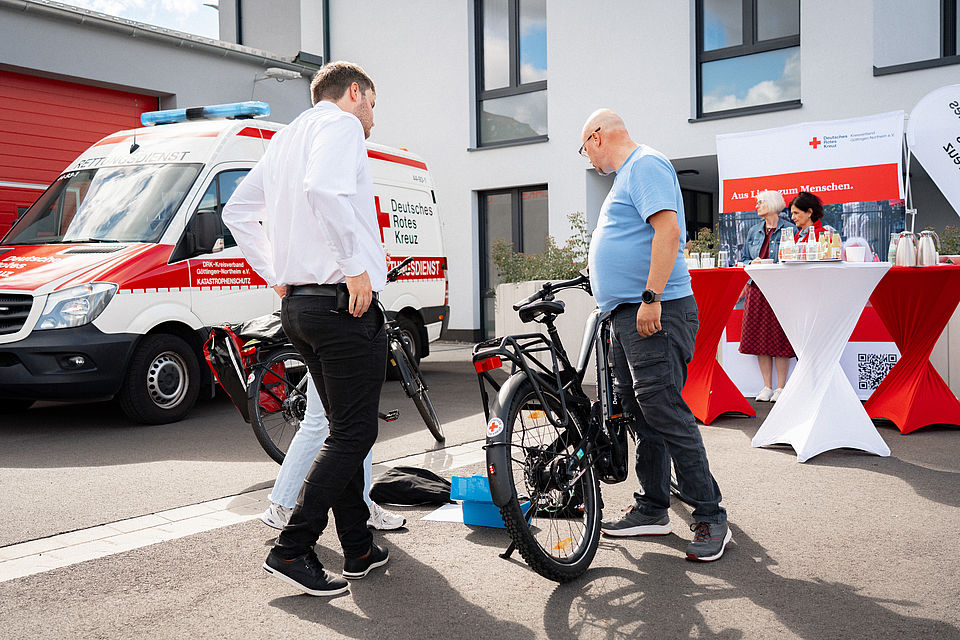 2026_08_11_Fahrradstaffel_Gründung.jpg Im Rahmen der Gründung der ersten Fahrradstaffel des DRK-Kreisverbands Göttingen-Northeim erklärt Stefan Trauth vom Förderer Bike2Future die Funktionen und Ausstattung eines der neuen E-Bikes. Die Vorstellung findet auf dem Gelände in der Zimmermannstraße 4 in Göttingen statt. Im Hintergrund sind ein Fahrzeug des Katastrophenschutzes sowie der Veranstaltungsbereich der offiziellen Fahrradübergabe zu sehen.