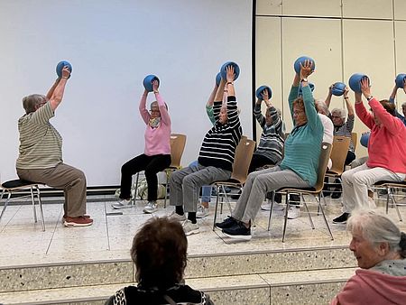 Mitglieder der Gymnastikgruppe des Ortsvereins Uslar sitzen auf Stühlen auf einer Bühne bei der Seniorenmesse in Uslar, alle halten einen Ball in die Luft, vorne ist Anleiterin Frau Andermann