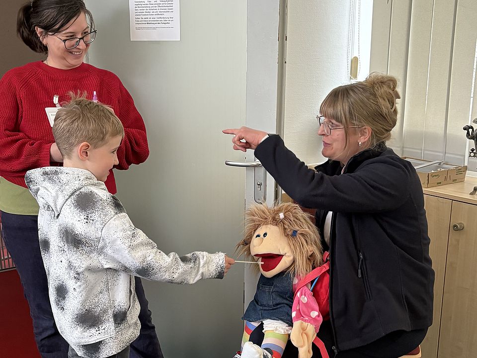 Kinder und Erwachsene der Evangelischen Familienbildungsstätte nehmen an einer interaktiven Lerneinheit mit einer DRK‑Puppe teil und erfahren beim Besuch des DRK-Kreisverbandes Göttingen‑Northeim mehr über den Ablauf einer Blutspende. Ein Kind benutzt ein Teststäbchen an der Blutspendepuppe, um einen Eisentest zu simulieren