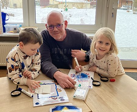An einem Holztisch in einem Gruppenraum in der DRK-Kita Scheden arbeiten zwei Kinder und eine erwachsene Begleitperson gemeinsam an einer Aufgabe. Vor ihnen liegen Lupen, Stifte, Papier und ein Schild in Form eines Polizeiabzeichens, das an die Paw‑Patrol‑Figur Chase erinnert. Die Kinder zeichnen auf Arbeitsblättern, während die erwachsene Person unterstützend die Materialien sortiert. Hinter ihnen zeigt ein großes Fenster den verschneiten Außenbereich der Kita. Auf dem Fenster hängt ein Paw‑Patrol‑Ballon. Die Szene gehört zur Spürnasen‑Station der Olympiade, bei der Kinder gemeinsam mit ihren männlichen Bezugspersonen Hinweise suchen und Detektivaufgaben lösen.