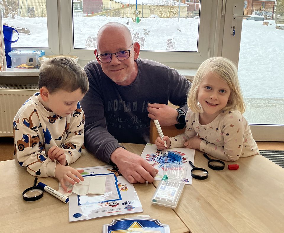 An einem Holztisch in einem Gruppenraum in der DRK-Kita Scheden arbeiten zwei Kinder und eine erwachsene Begleitperson gemeinsam an einer Aufgabe. Vor ihnen liegen Lupen, Stifte, Papier und ein Schild in Form eines Polizeiabzeichens, das an die Paw‑Patrol‑Figur Chase erinnert. Die Kinder zeichnen auf Arbeitsblättern, während die erwachsene Person unterstützend die Materialien sortiert. Hinter ihnen zeigt ein großes Fenster den verschneiten Außenbereich der Kita. Auf dem Fenster hängt ein Paw‑Patrol‑Ballon. Die Szene gehört zur Spürnasen‑Station der Olympiade, bei der Kinder gemeinsam mit ihren männlichen Bezugspersonen Hinweise suchen und Detektivaufgaben lösen.