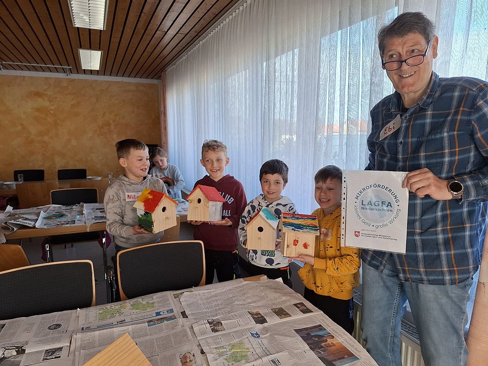 Kinder beim Nistkästen bauen für den Walderlebnistag des DRK-Ortsvereins Harste in einer Gruppe im Wald, in der Hand halten sie ihre gebauten Nistkästen, zudem hält ein Ehrenamtlicher einen Zettel mit dem Logo des Spendengebers