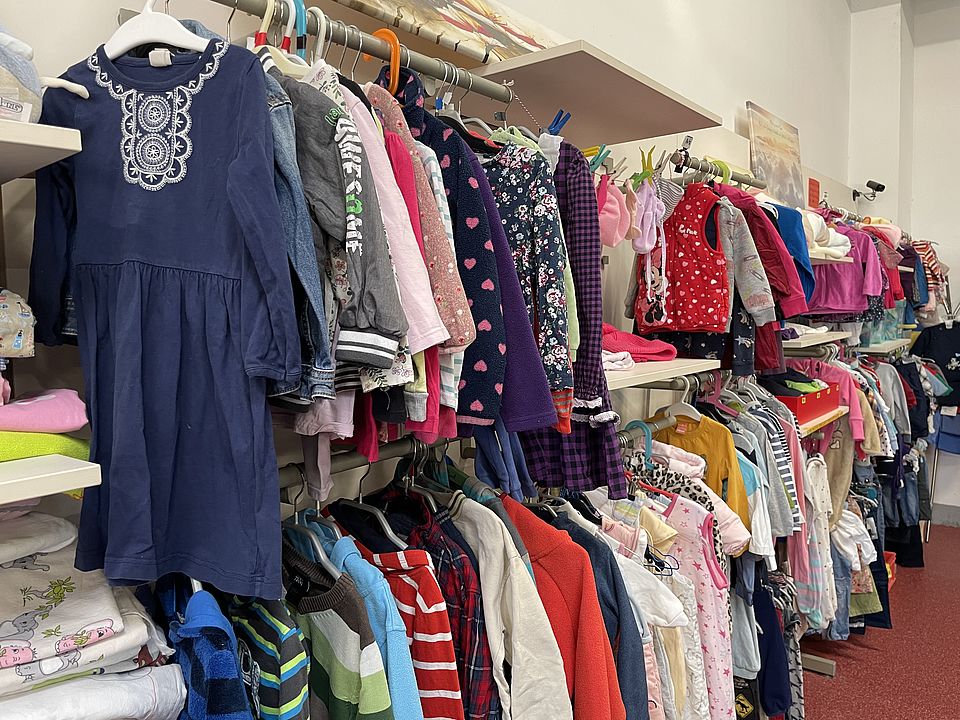 Kleiderstangen, die mit verschiedenen Kleidungsstücken bestückt sind und Regale, die mit Ware bepackt sind, sind an einer der Wände des Raumes zu sehen.