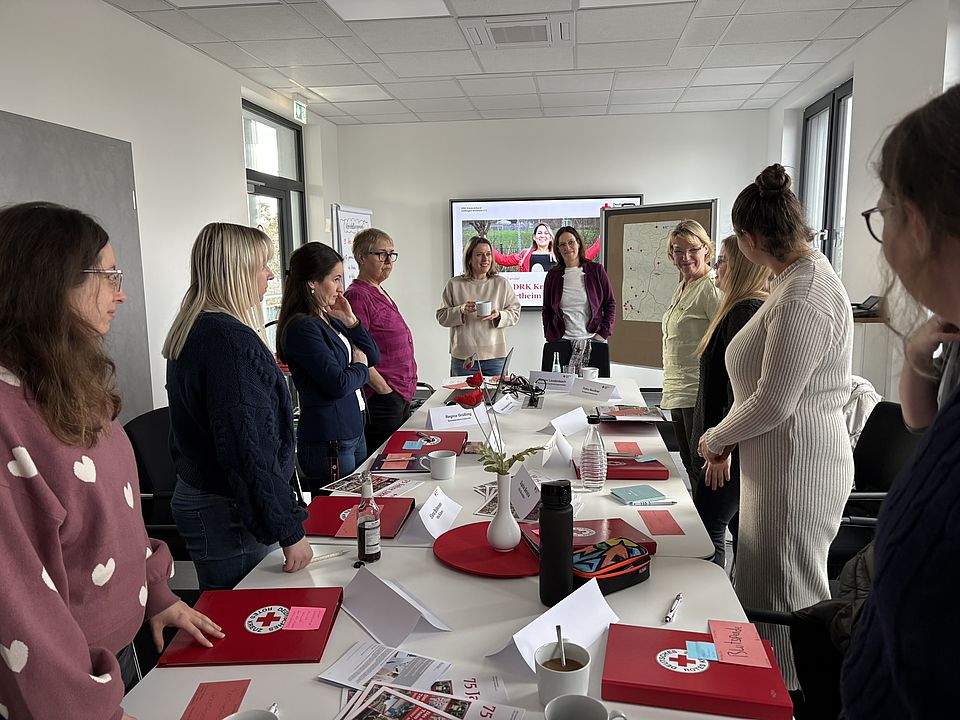Willkommenstag des DRK-Kreisverbandes Göttingen‑Northeim: Teilnehmende stehen an einem Konferenztisch, während vorne Präsentationsmaterial sowie eine Standortkarte erläutert werden. Auf dem Tisch liegen Unterlagen und DRK‑Materialien bereit