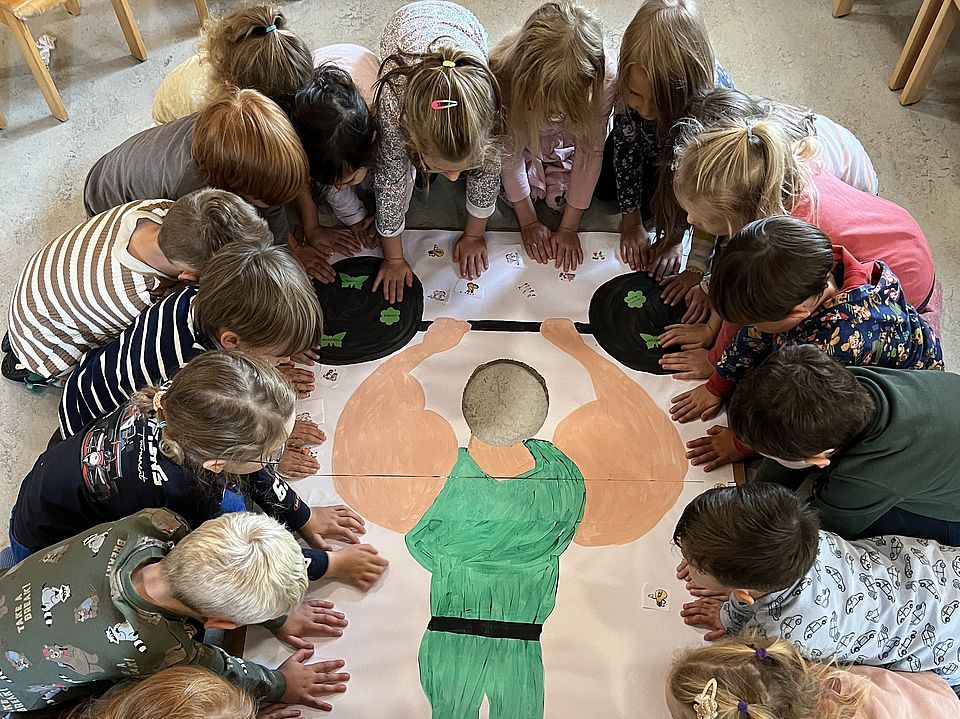 2026_09_21_Kindertag_Kita_Volkmarshausen__4_.jpeg Kinder der DRK-Kita Hedemünden im Kreis auf dem Boden, in der Mitte ein Bild von einem starken Gewichtheber