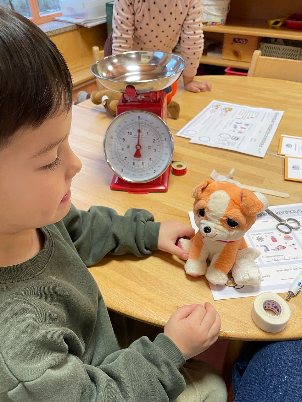 2025_11_24_Teddybärkrankenhaus__5 Ein Kind sitzt an einem Tisch auf dem eine Waage steht und hat ein Kuscheltier in der Hand, einen kleinen Hund. Im Hintergrund ist ein zweites Kind zu erahnen