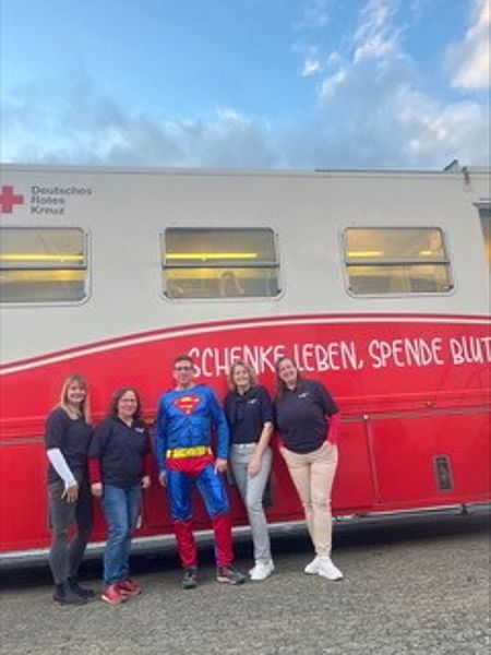 Vier Frauen des DRK-OV Hollenstedt und ein junger Mann, der als Superman verkleidet ist, stehen vor dem Blutspendemobil-Bus