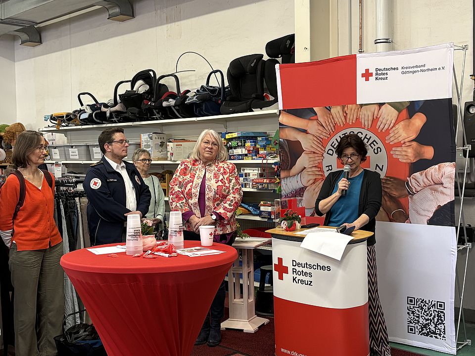 2026_03_03_Rede_Petra_Reußner_Spnedenzentrum_Einweihung.jpg DRK-Kreisgeschäftsführerin Petra Reußner spricht am DRK‑Redepult im Spendenzentrum Göttingen, umgeben von Gästen der Einweihung. Hinter dem Redepult steht ein großes DRK‑Banner mit dem Motiv vieler übereinandergelegter Hände.