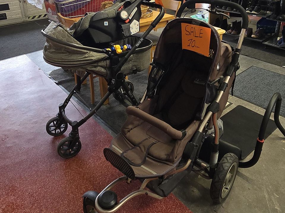Innenraum des DRK-Spendenzentrums. Im Vordergrund stehen zwei gebrauchte Kinderwagen; einer davon hat ein orangefarbenes Schild mit der Aufschrift ‚Sale‘. Hinter den Kinderwagen steht ein Einkaufswagen, gefüllt mit mehreren großen, bunten Plastikverpackungen. Im Hintergrund befinden sich Regale mit Haushaltsgegenständen und zwei Personen, die sich dort umsehen. Der Raum ist groß und industriell mit sichtbaren Lüftungsschächten und einer Glasfront an der linken Seite.