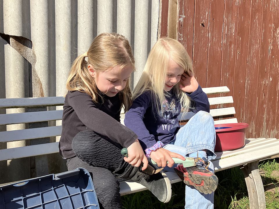 Reitstallausflug der DRK-Kita Scheden: Zwei Kinder sitzen auf einer Bank und üben das Hufeauskratzen eines Pferdes an ihren eigenen Schuhen