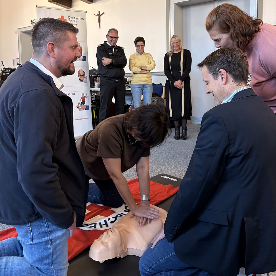 2026_09_21_Woche_der_Wiederbelebung__2_.jpg Mehrere Gäste der Veranstaltung sowie DRK-Kreisgeschäftsführerin Petra Reußner probieren die Herz-Lungen-Wiederbelebung an einem Übungsphantom aus. Ehrenamtliche des Deutschen Roten Kreuzes begleiten die Übung und geben praktische Hinweise. Im Hintergrund verfolgen weitere Teilnehmende die Demonstration