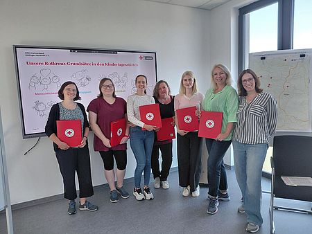 Neue Mitarbeitende in DRK-Kitas mit roten DRK-Mappen stehen vor einem Bildschirm mit einer Präsentation vom Roten Kreuz, gemeinsam mit DRK-Kreisverbands-Personalleitung Marion Laudenbach