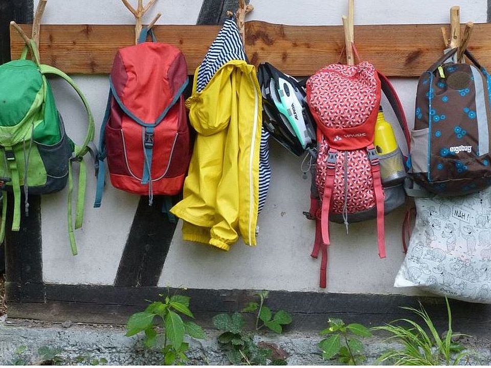 Mehrere Kinderrucksäcke in bunten Farben hängen an einer Holzgarderobe an der Außenwand des Kindergarten Häuschens im Wald