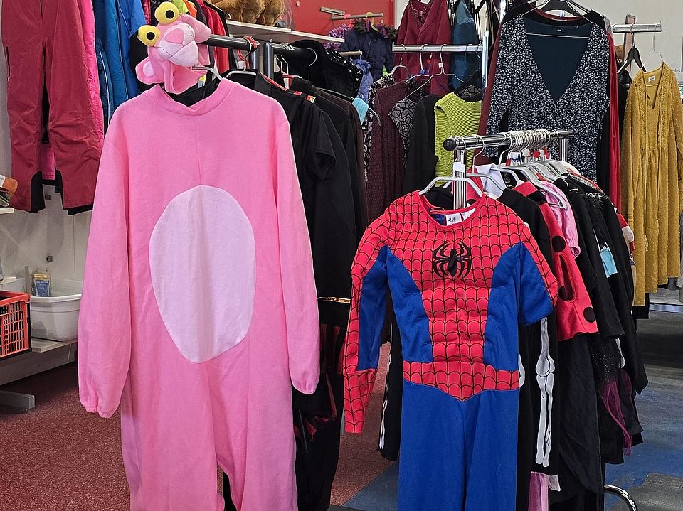 Innenraum des DRK-Spendenzentrums in Göttingen. Im Vordergrund hängen zwei Kostüme auf Kleiderständern: links ein großes pinkes Tierkostüm mit rundem weißem Bauch und Kapuze mit Augen; rechts ein blau‑rotes Kinderkostüm im Superhelden‑Stil. Dahinter stehen Kleiderständer mit verschiedenfarbiger Gebrauchtkleidung wie Jacken, Kleidern und Pullovern. Der Raum hat hohe Decken mit sichtbaren Metallträgern.