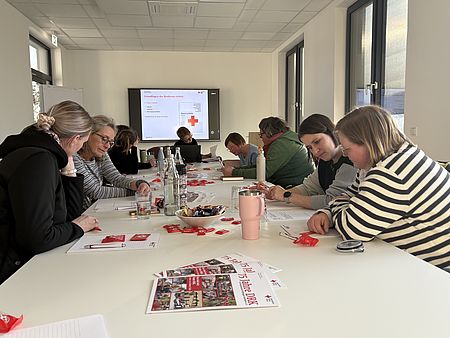 in heller Schulungsraum, in dem mehrere Personen an einem langen Konferenztisch sitzen und an einer Einführungsveranstaltung für neue Schulbegleiterinnen des FUD im DRK‑Kreisverband Göttingen‑Northeim e. V. teilnehmen. Auf einem großen Bildschirm an der Stirnseite des Raums ist eine Präsentationsfolie mit Informationen des Deutschen Roten Kreuzes zu sehen. Die Teilnehmenden schreiben Notizen, betrachten Unterlagen oder folgen der Präsentation. Auf dem Tisch liegen Schulungsmaterialien, DRK‑Informationsbroschüren, Schreibutensilien sowie zahlreiche kleine rote Give‑aways. Außerdem stehen Getränke, Wasserflaschen, Tassen und eine Schale mit Süßigkeiten bereit. Die Schulung wird von Alexandra Jäger geleitet. Durch die großen Fenster fällt viel Tageslicht in den Raum.