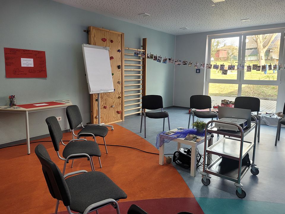 Vorbereiteter Raum für das Elterncafé in der Kita: Stuhlkreis, Flipchart, Tisch mit Materialien und eine Fotoleine mit Bildern aus dem Kita‑Alltag. Durch die Fenster ist der Außenbereich zu sehen
