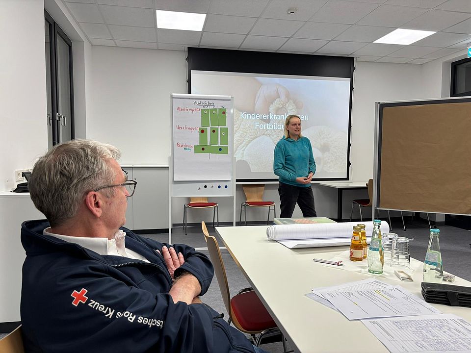 2026_01_29_Ausbilder_Fortbildung_Erste_Hilfe__1_.jpg In einem Schulungsraum des DRK-Kreisverbandes Göttingen‑Northeim steht Referentin Tessa Lehmann vor einer Leinwand, auf der eine Präsentationsfolie mit dem Titel „Kindererkrankungen – Fortbildung“ angezeigt wird. Neben ihr befindet sich ein Flipchart mit einer grafischen Darstellung. Im Vordergrund sitzt Bereichsleiter Markus-Schiffer in einer dunkelblauen DRK‑Jacke und hört der Präsentation zu. Auf den Tischen liegen Unterlagen, Schreibmaterialien, Getränke und Schulungsmaterial. Die Szene zeigt einen Moment der praxisnahen Erste-Hilfe-Fortbildung für Ausbilder*innen, die von Markus Schiffer, Tessa Lehmann und Niklas Benndorf gestaltet wird.