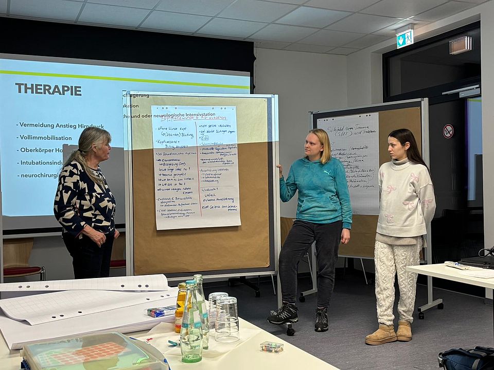2026_01_29_Ausbilder_Fortbildung_Erste_Hilfe__4_.jpg In einem Schulungsraum des DRK-Kreisverbands Göttingen‑Northeim stehen Tessa Lehmann und zwei Teilnehmende vorne im Raum und präsentieren Ergebnisse einer Gruppenarbeit. Vor ihnen steht ein großes Moderationsboard mit mehreren handschriftlich ausgefüllten Papierbögen, auf denen Symptome, Maßnahmen und wichtige Stichpunkte notiert sind. Links daneben wird auf einer Leinwand eine Präsentationsfolie mit dem Titel „Therapie“ angezeigt. Im Vordergrund des Bildes stehen auf einem Tisch mehrere Getränke, Unterlagen, Schreibmaterialien und Schulungsmappen. Die Szene zeigt einen interaktiven Teil der praxisnahen Erste‑Hilfe‑Fortbildung für Ausbilder*innen, die von Markus Schiffer, Tessa Lehmann und Niklas Benndorf gestaltet wird.