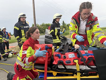 Rettungsdienstübung Uslar: drei Auszubildende des DRK-Rettungsdienstes in rot-gelber Uniform kümmern sich um Patientin auf einer Liege vor dem Auto, im Hintergrund Feuerwehr
